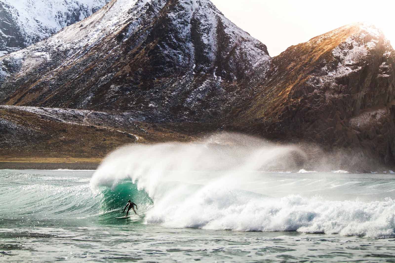 Surfer i en grønn bølge med sjøsprøyt og snødekte fjell i bakgrunnen